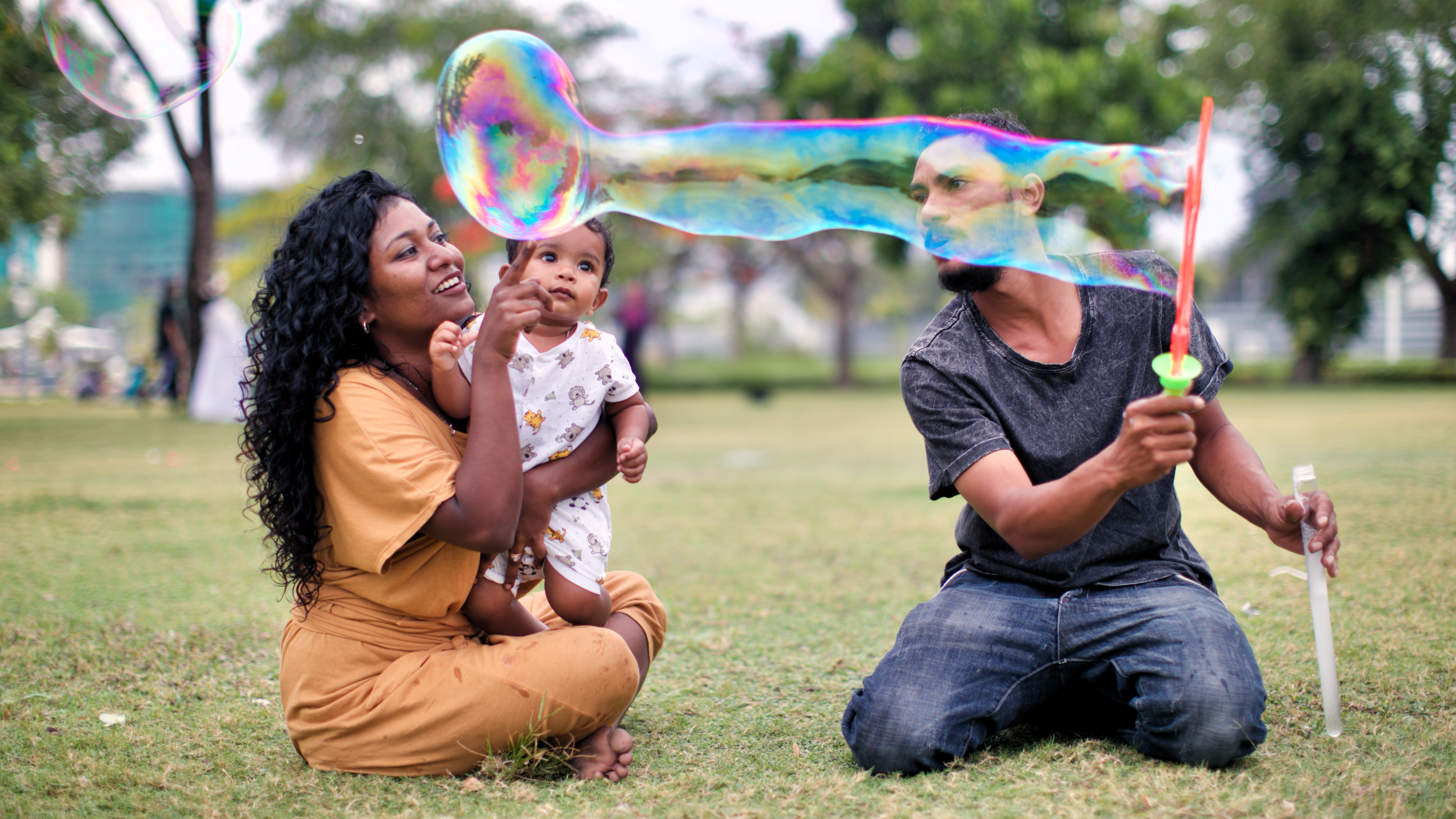 A family, mom holds a baby boy and the dad is blowing bubbles in the park