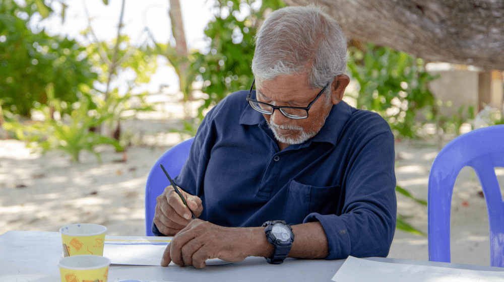 An elderly man paints on a white canvas by the beach.