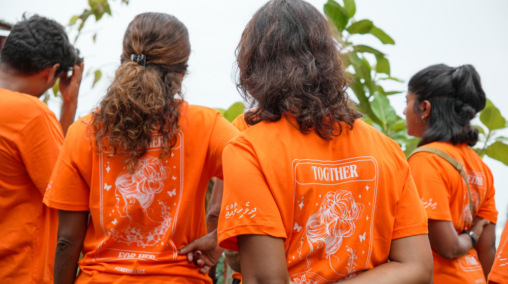 Four women stand with their backs faced, wearing graphic orange t-shirts that read "Together For Her".